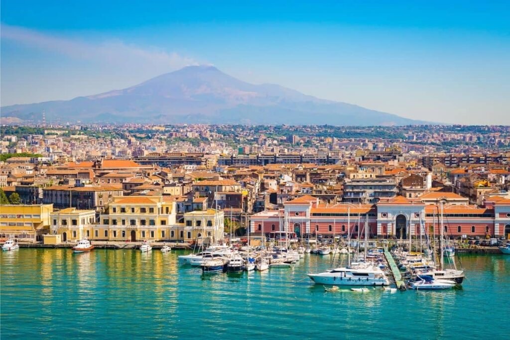 View of Catania with the blue colours of the bay in the foreground, buildings of the city in the middle of the picture and Mt Etna looming over the city in the background