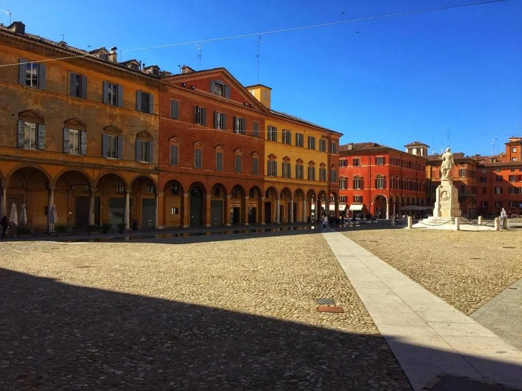 Piazza in Modena with multicoloured buildings in the background and a piazza in the foreground with a fountain to the right of the image