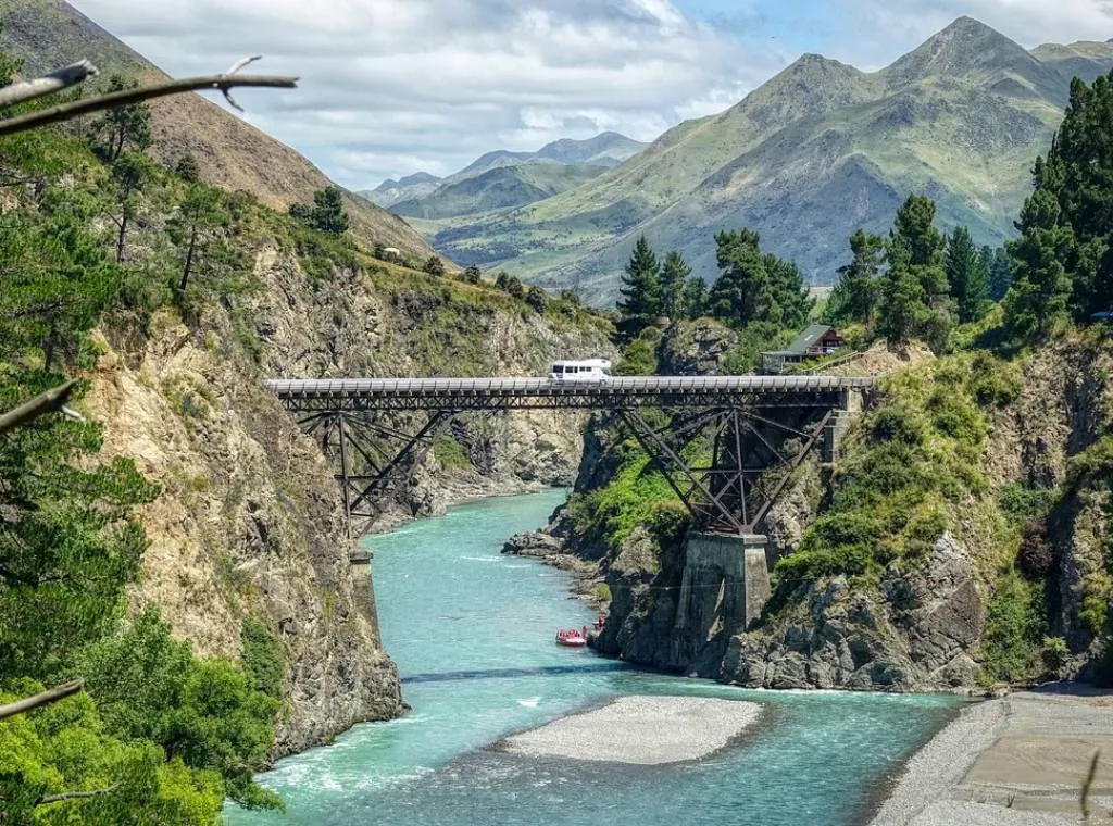 a wooden bridge in hanmer springs with blue water underneath and mountains in the background