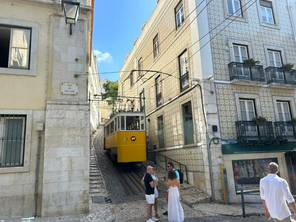 a yellow tram in Lisbon in between two buildings at the bottom of a hill